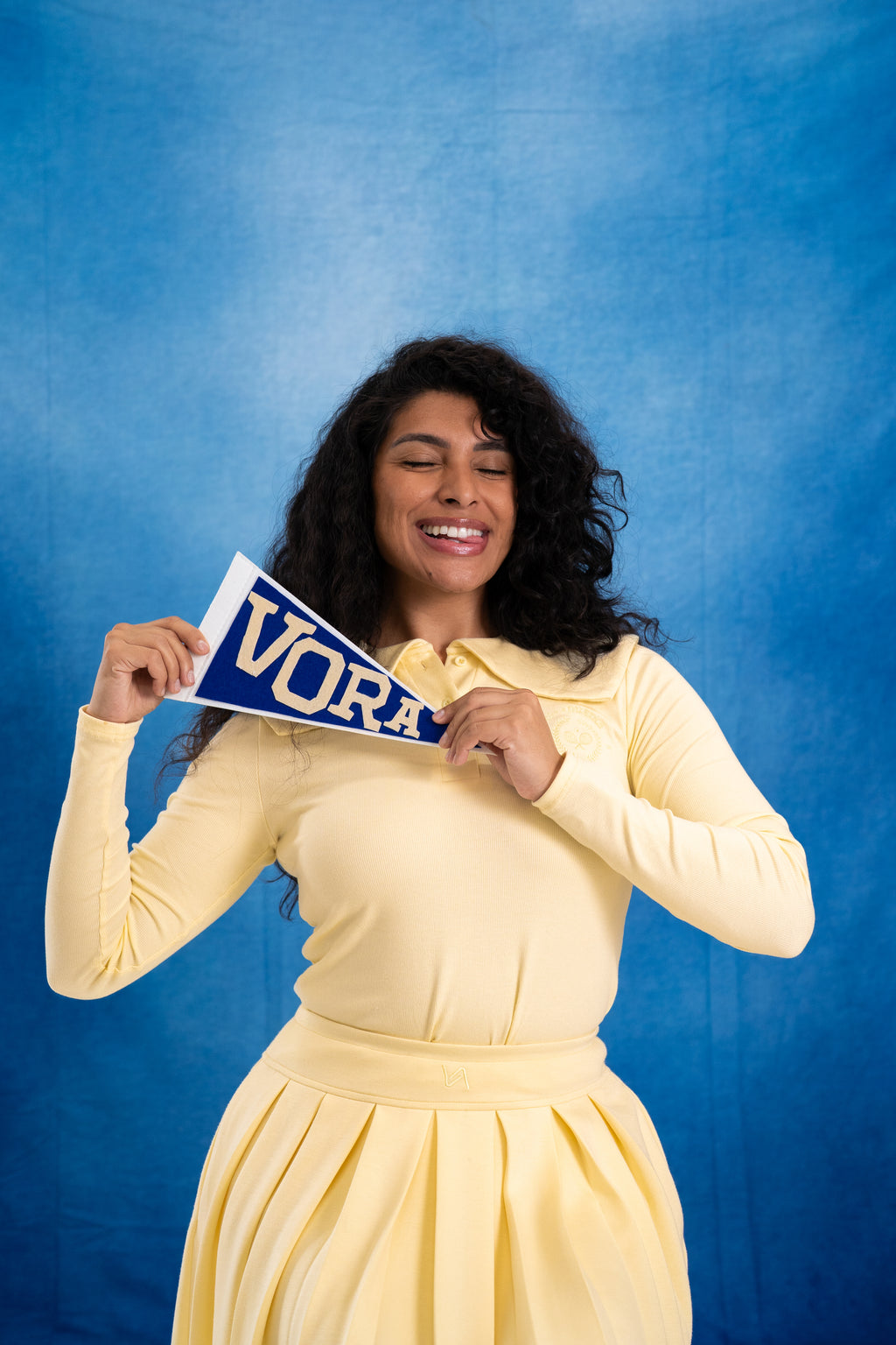 Woman in a yellow set holding up a blue pennant flag with 'VORA' on it against a blue background