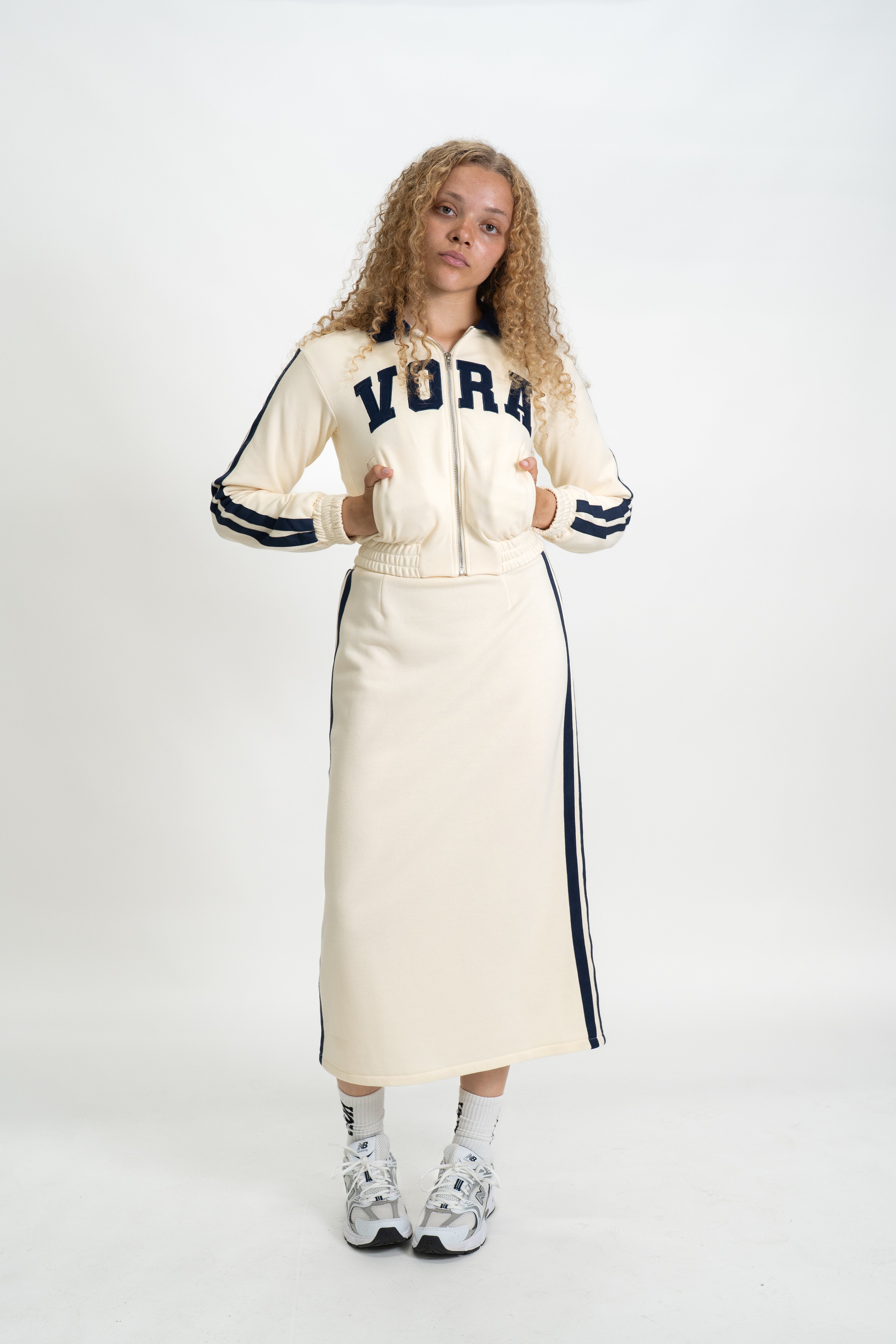 Woman wearing a cream-colored jacket with 'VORA' branding and a long skirt on a white background
