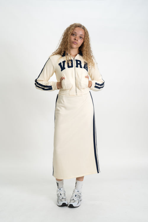 Woman wearing a cream-colored jacket with 'VORA' branding and a long skirt on a white background