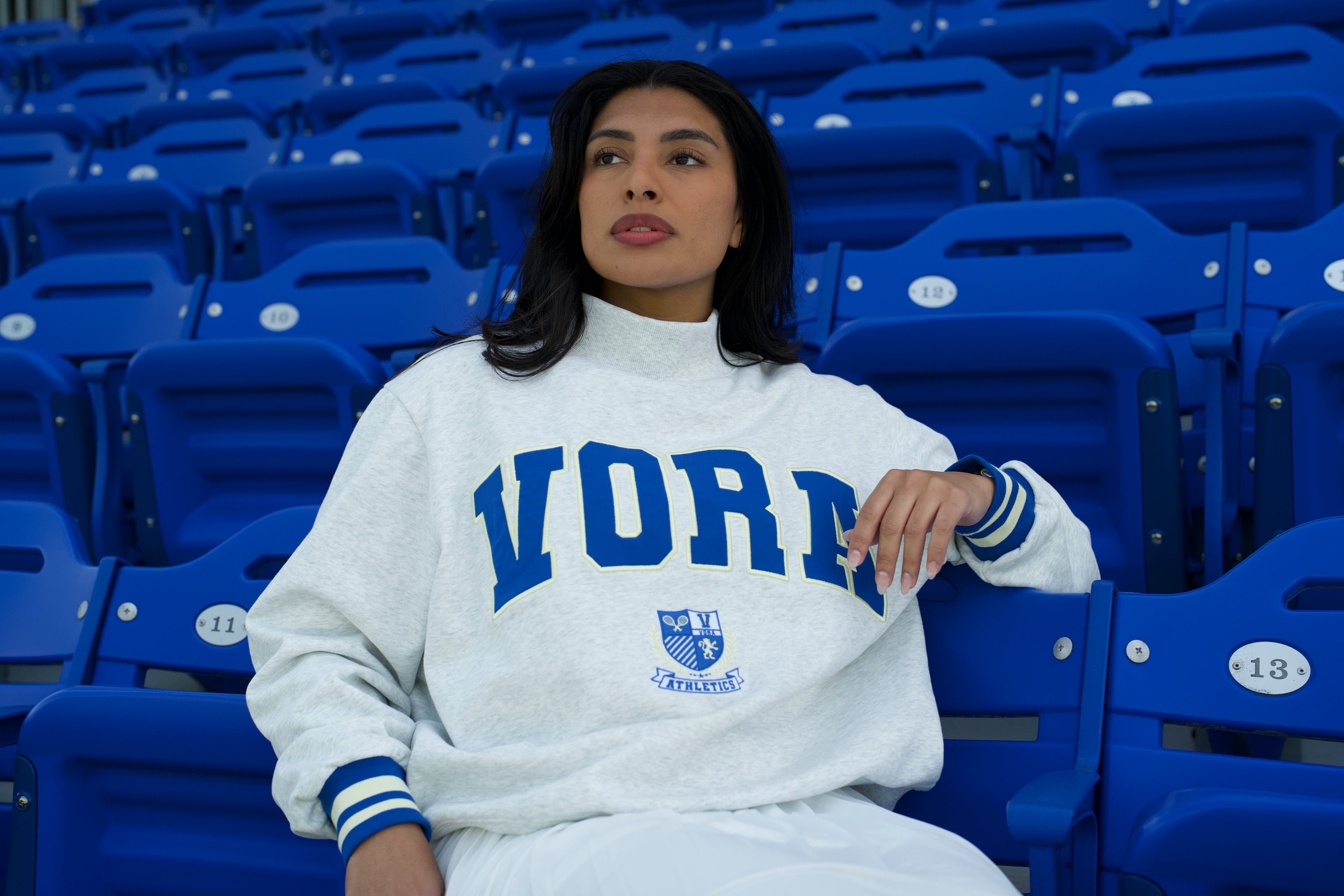 Person wearing a light grey sweatshirt with blue text sitting in a stadium with blue seats.