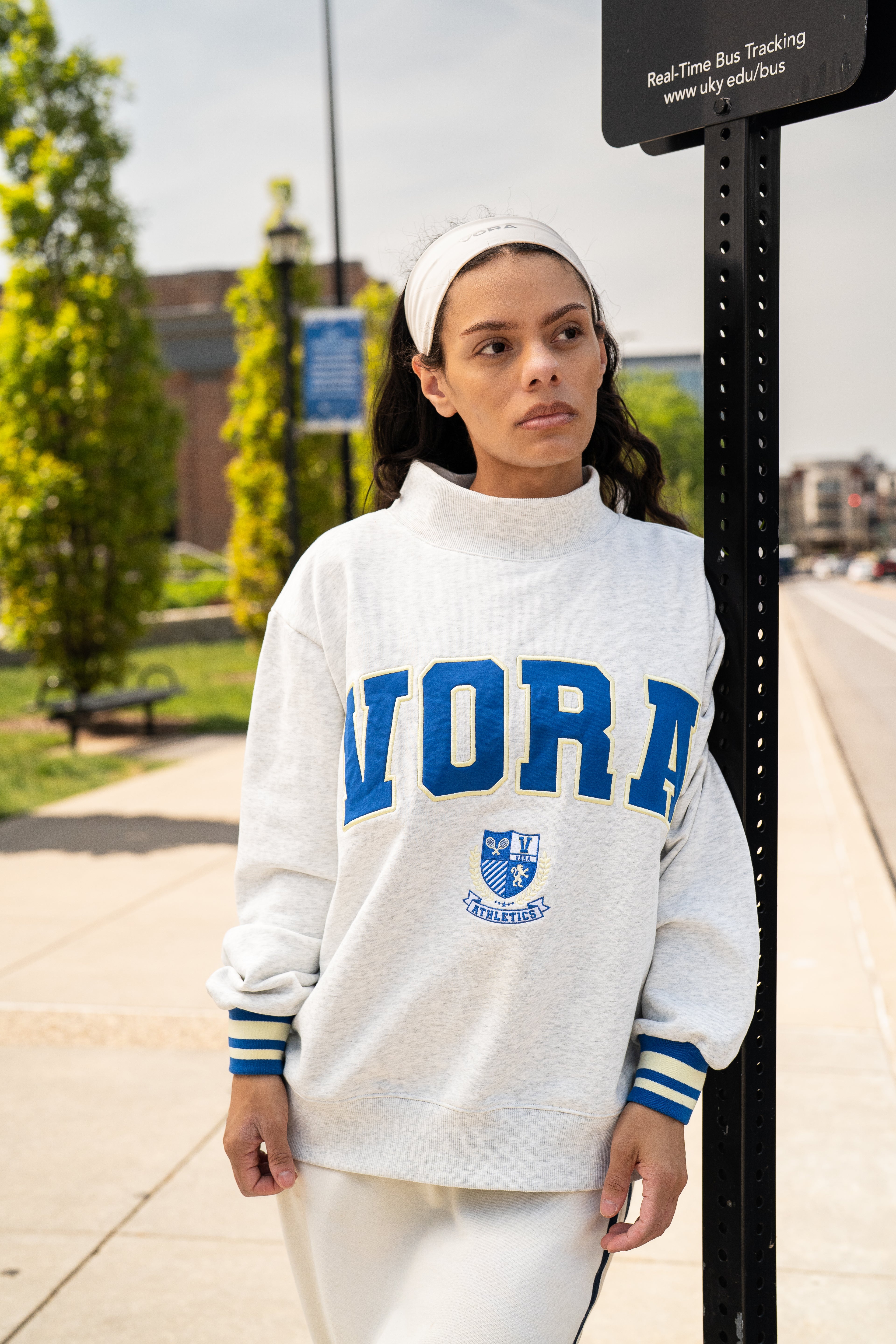 Woman standing net to bus stop in a light grey sweatshirt with VORA written across the front in cobalt blue. 
